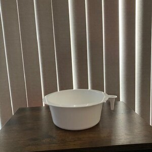 May include: A white plastic bowl with a smooth, rounded interior and exterior surface. The bowl features a small, vertical handle extending from one side, designed for easy gripping. The construction is simple, featuring a wide, open top rim.