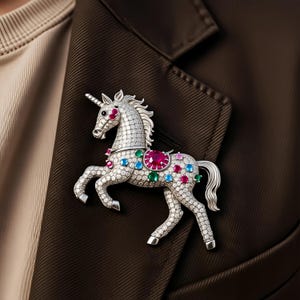 May include: A silver-toned unicorn brooch with a horn and flowing mane. The brooch is encrusted with clear and colorful gemstones, including red, blue, and green. The brooch is attached to a brown blazer.