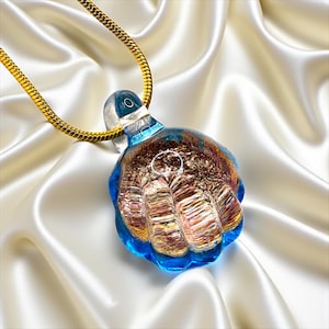 May include: A unique glass pendant necklace featuring a fist-shaped design. The pendant has a blue, brown, and gold color scheme and is suspended from a gold chain. The necklace is displayed on a white satin background.