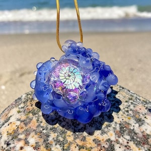 May include: A pendant featuring a central iridescent ammonite fossil encased in translucent blue glass with a textured, bubbly surface. The piece is suspended from a thin gold-toned snake chain, showcasing a swirling shell pattern with purple and silver highlights.