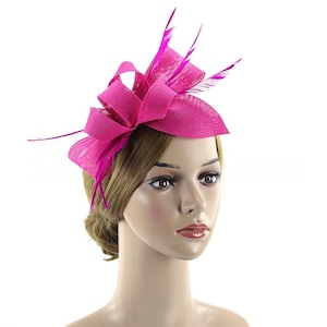 Op de afbeelding: Een felroze fascinator met een strik en veermotieven. Het hoofddeksel is gemaakt van een geweven materiaal en heeft een grote strik bovenop met verschillende lange, dunne veren die omhoog steken. De fascinator is ontworpen om op het hoofd te worden gedragen.
