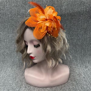May include: An orange floral headpiece with a mesh overlay, feathers, and beaded accents. The headpiece is attached to a mannequin head with wavy blonde hair. The headpiece is a vibrant accessory.