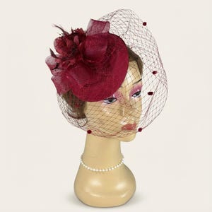 May include: A burgundy fascinator with a veil, displayed on a mannequin head. The hat features a round base, adorned with fabric flowers and feathers. A net veil with small, round accents drapes over the face, and a pearl necklace is around the neck.