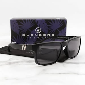 May include: Black sunglasses with a rectangular frame and dark lenses. The sunglasses have a silver stripe on the temples and are resting on a black case. A box with the brand name "Blenders Eyewear" is in the background.