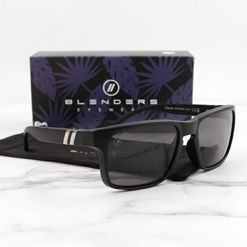 May include: Black sunglasses with a rectangular frame and dark lenses. The sunglasses have a silver stripe on the temples and are resting on a black case. A box with the brand name "Blenders Eyewear" is in the background.