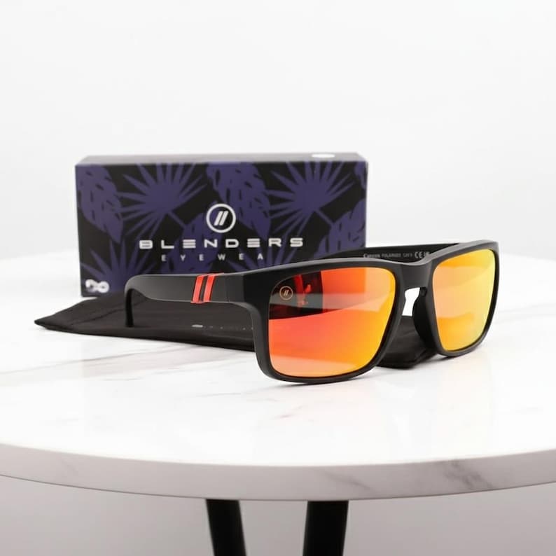May include: Black sunglasses with orange mirrored lenses and red stripes on the temples. The sunglasses are displayed on a black cloth with a box in the background that says "Blenders Eyewear".