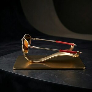 May include: A pair of sunglasses with orange lenses and silver frames, accented with red temple tips, rests on a gold-colored display stand. The stand has a rectangular base and a curved, wave-like design.