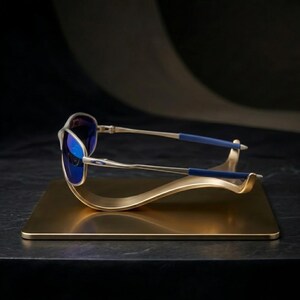May include: Silver-framed sunglasses with blue lenses rest on a gold-colored display stand. The sunglasses have blue accents on the temples. The stand is a rectangular base with a curved, wave-like design.