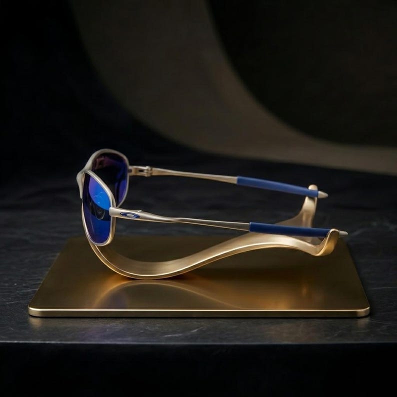 May include: Silver-framed sunglasses with blue lenses rest on a gold-colored display stand. The sunglasses have blue accents on the temples. The stand is a rectangular base with a curved, wave-like design.