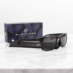 May include: Black sunglasses with dark lenses and a matte finish, resting on a black pouch. A box with the text "BLENDERS EYEWEAR" and a purple leaf pattern is in the background. The sunglasses have a rectangular frame.