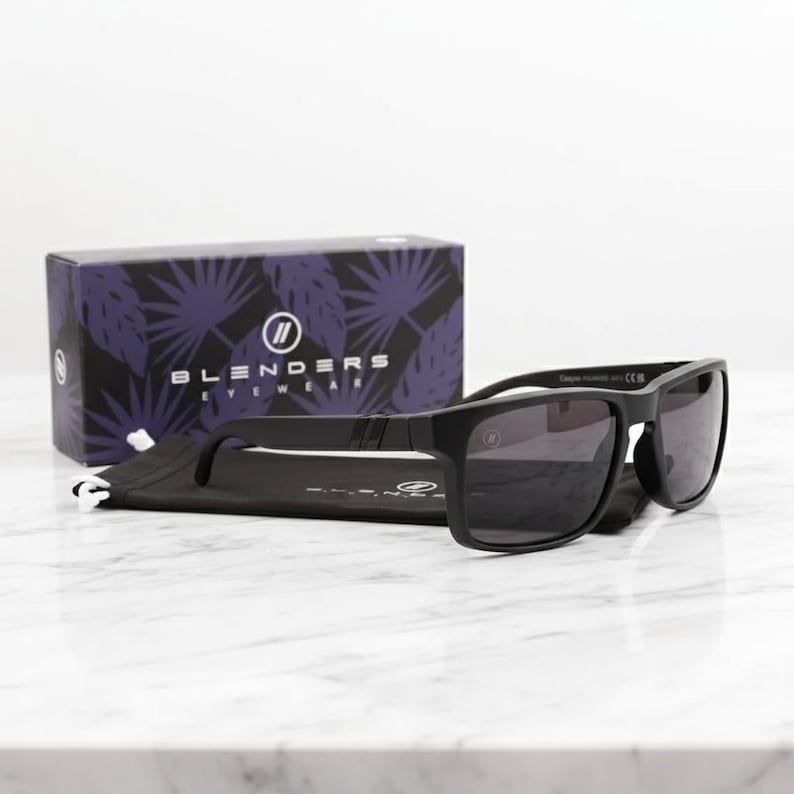 May include: Black sunglasses with dark lenses and a matte finish, resting on a black pouch. A box with the text "BLENDERS EYEWEAR" and a purple leaf pattern is in the background. The sunglasses have a rectangular frame.