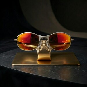 May include: Silver-framed sunglasses with vibrant red-orange lenses are displayed on a gold-colored stand. The sunglasses have a sleek, modern design, and the stand is rectangular with a curved support.