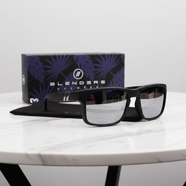 May include: Black sunglasses with silver mirrored lenses rest on a black case, in front of a box with the text "BLENDERS EYEWEAR". The box has a purple and white leaf pattern. The sunglasses are on a white marble surface.