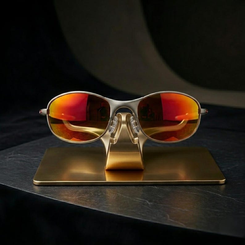 May include: Silver-framed sunglasses with vibrant red-orange lenses are displayed on a gold-colored stand. The sunglasses have a sleek, modern design, and the stand is rectangular with a curved support.