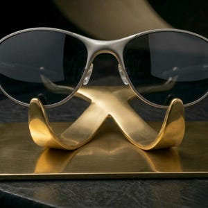 Op de afbeelding: Zonnebril met zilveren montuur en donkere glazen rust op een goudkleurige metalen displaystandaard. De standaard heeft een gestileerde X-vorm met gebogen armen en staat op een rechthoekige basis. De zonnebril is het middelpunt.