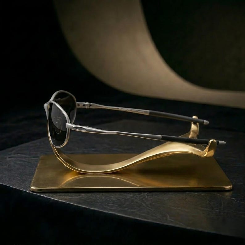 Op de afbeelding: Zilverkleurige zonnebril met donkere glazen rust op een goudkleurige displaystandaard. De standaard heeft een rechthoekige basis en een gebogen, golfachtig ontwerp om de bril te houden. De achtergrond is donker.