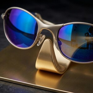 May include: Close-up of silver-framed sunglasses with blue mirrored lenses, resting on a gold-colored display stand. The lenses reflect a vibrant blue and the frame has a matte finish. The stand is also gold and has a curved design.