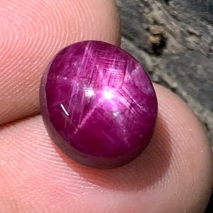 Peut inclure: Une pierre précieuse polie, de forme ovale, rouge foncé, avec un motif en forme d'étoile. La gemme a une surface lisse et réfléchissante. L'effet d'étoile est créé par la réflexion de la lumière sur les inclusions de la pierre.