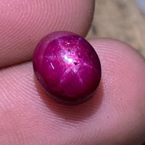 May include: A polished, oval-shaped ruby gemstone. The gem is a deep, saturated pinkish-red color with a star-like effect visible on its surface. The stone is smooth and reflects light, showcasing its natural beauty.