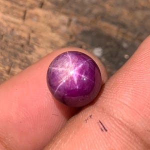 May include: A round, cabochon-cut star ruby gemstone with a deep magenta-purple hue. The stone features a distinct six-rayed asterism, or star effect, centered on its polished, smooth, and domed surface, reflecting light in a bright, sharp pattern.