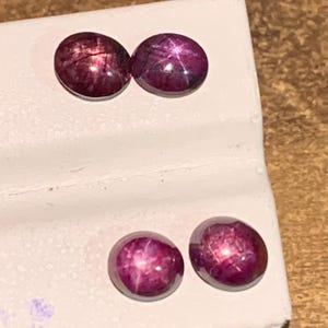 May include: Four round, cabochon-cut star rubies with a deep magenta-red hue. Each gemstone displays a distinct six-rayed star effect, known as asterism, visible on the smooth, polished surface of the stones when light hits the center point.