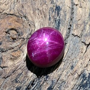 Puede incluir: Una piedra preciosa cabujón pulida de forma ovalada en un color rojo magenta profundo y vibrante. La superficie de la piedra muestra un patrón de estrella de seis rayos, conocido como asterismo, que refleja la luz sobre la parte superior redondeada.