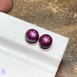 May include: Two round, cabochon-cut star rubies with a deep magenta-red hue. Each gemstone displays a distinct six-rayed asterism effect, characterized by a bright, intersecting light pattern visible on the smooth, polished, domed surfaces.