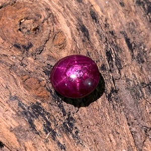 May include: A round, polished ruby gemstone with a star effect, resting on a textured, brown wooden surface. The gemstone is a deep, vibrant red with a star-like pattern radiating from the center.