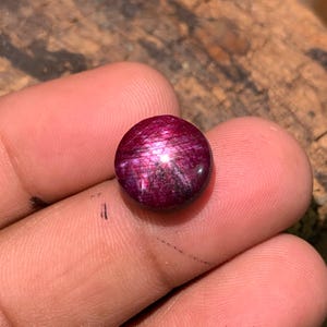 May include: A round, cabochon-cut star ruby gemstone with a deep magenta-red color. The stone displays a distinct six-rayed star effect, known as asterism, which appears as a bright white light reflection crossing the center of the surface.