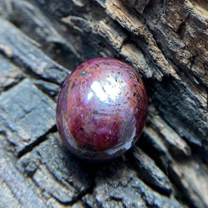 May include: A polished, oval-shaped ruby gemstone featuring a deep magenta and dark red color palette with visible natural inclusions and dark speckles throughout the surface, reflecting light with a smooth, rounded, and glossy finish.