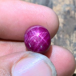 May include: A round, cabochon-cut star ruby gemstone with a deep magenta-red color. The stone displays a distinct six-rayed star effect, known as asterism, visible across its polished, domed surface under a bright light source.