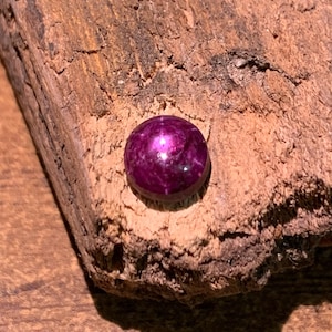 May include: A round, cabochon-cut gemstone with a deep magenta and purple hue. The stone features a smooth, polished surface with a subtle star-like reflection visible in the center, resting on a piece of rough, textured, light brown bark.