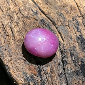 May include: A polished, oval-shaped, pink gemstone with a star-like pattern. The gem is set against a textured, weathered, brown wooden surface. The star effect is visible across the surface of the stone.