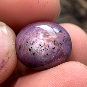 May include: A round, cabochon-cut gemstone with a deep purple and magenta hue. The surface displays a visible star-like optical phenomenon, known as asterism, with intersecting light rays and scattered dark mineral inclusions throughout the stone.