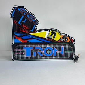 May include: A Tron arcade marquee sign featuring a black background with a grid pattern, the word TRON in blue and pink lettering, and a graphic of a yellow and orange light cycle. The sign includes a black power cord with a USB connector.