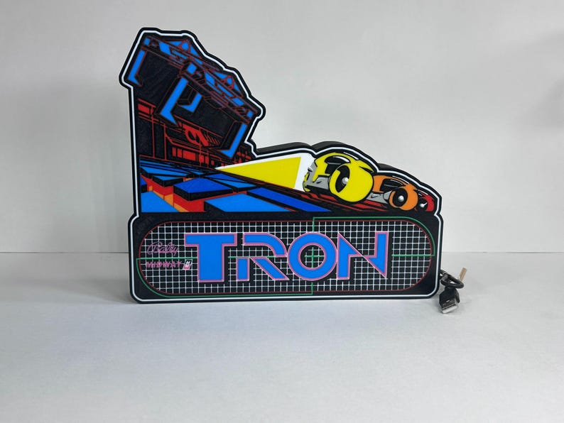 May include: A Tron arcade marquee sign featuring a black background with a grid pattern, the word TRON in blue and pink lettering, and a graphic of a yellow and orange light cycle. The sign includes a black power cord with a USB connector.