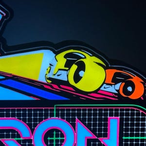 May include: A neon-style sign featuring a vibrant graphic of two futuristic vehicles in yellow and orange, casting a bright yellow light beam. Below the vehicles, the word TRON is displayed in large, bold blue letters against a black grid background.