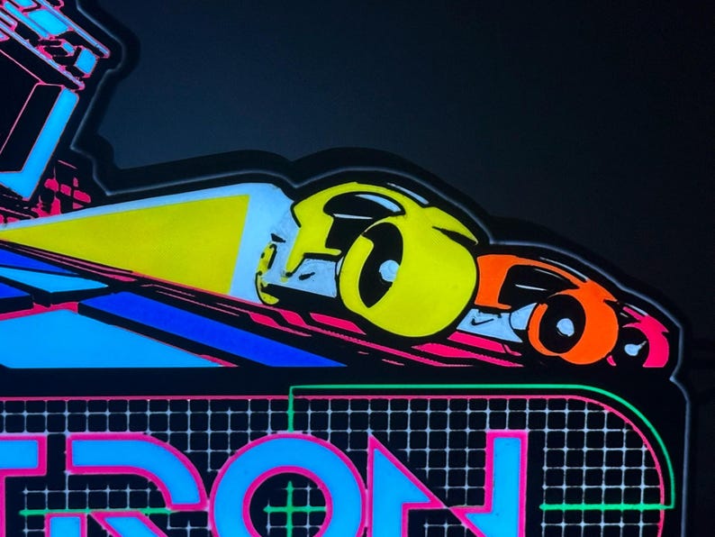 May include: A neon-style sign featuring a vibrant graphic of two futuristic vehicles in yellow and orange, casting a bright yellow light beam. Below the vehicles, the word TRON is displayed in large, bold blue letters against a black grid background.
