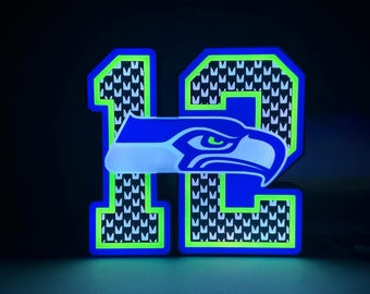 May include: Illuminated blue and green number 12 with a Seattle Seahawks logo. The number has a black and white pattern inside. The Seahawks logo is a stylized eagle head in blue, white, and green.