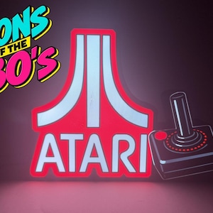 May include: A glowing neon-style sign featuring the white and red Atari logo with the text ICONS OF THE 80'S in blue and pink lettering above it. To the right of the logo is a black joystick controller with a single red button and red accents.