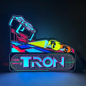 May include: A glowing LED sign featuring the word TRON in bright cyan and pink letters over a grid pattern. Above the text, a stylised graphic shows two vehicles racing on a track with a yellow light beam, all set against a dark background.