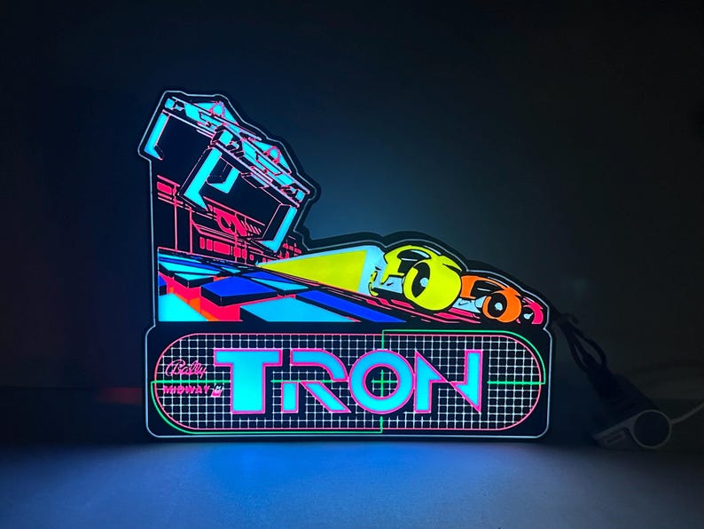 May include: A glowing LED sign featuring the word TRON in bright cyan and pink letters over a grid pattern. Above the text, a stylised graphic shows two vehicles racing on a track with a yellow light beam, all set against a dark background.