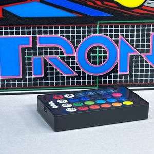 May include: A rectangular black remote control with the brand name onn. printed in white. The remote features a grid of twenty-four circular buttons in various colours and functions, positioned in front of a graphic panel displaying the word TRON.