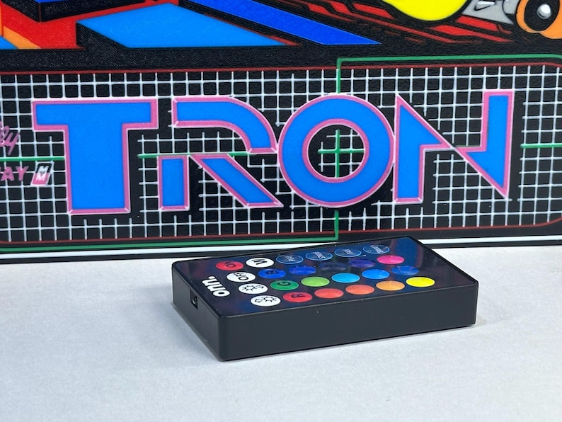 May include: A rectangular black remote control with the brand name onn. printed in white. The remote features a grid of twenty-four circular buttons in various colours and functions, positioned in front of a graphic panel displaying the word TRON.