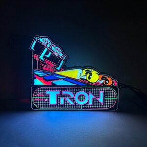 May include: A glowing Tron arcade marquee sign featuring a vibrant neon aesthetic. The design displays the word TRON in bold, cyan-outlined lettering over a dark grid pattern, with a stylised yellow and orange vehicle graphic above the text.