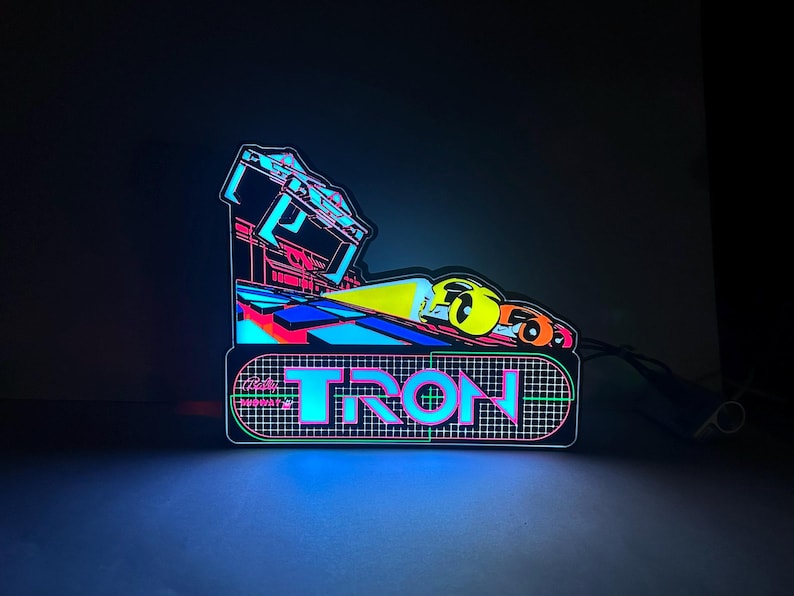 May include: A glowing Tron arcade marquee sign featuring a vibrant neon aesthetic. The design displays the word TRON in bold, cyan-outlined lettering over a dark grid pattern, with a stylised yellow and orange vehicle graphic above the text.