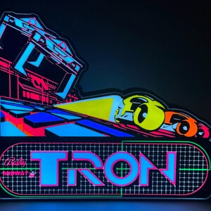May include: A neon-style light sign featuring the word TRON in bright blue letters over a grid pattern. Above the text, a yellow light beam shines from a vehicle on a blue and black geometric surface, with Bally Midway branding visible.