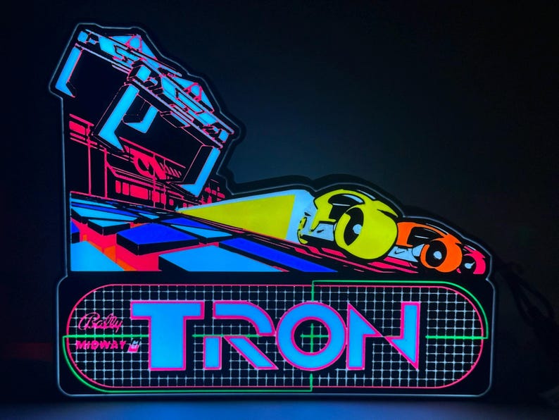 May include: A neon-style light sign featuring the word TRON in bright blue letters over a grid pattern. Above the text, a yellow light beam shines from a vehicle on a blue and black geometric surface, with Bally Midway branding visible.