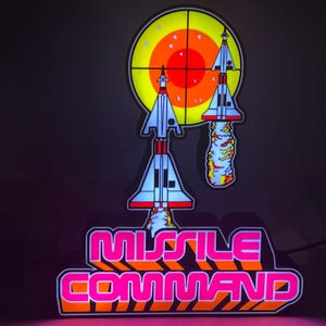 May include: A neon sign featuring the words "MISSILE COMMAND" in pink and orange, with a retro font. Above the text are two stylized rockets and a target. The sign has a black background.