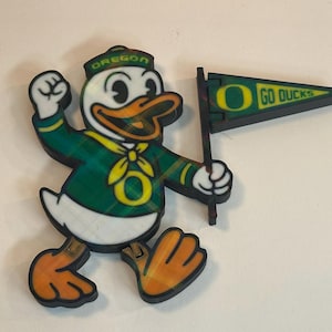 May include: A wooden cutout of the Oregon Ducks mascot, a duck wearing a green and yellow plaid outfit and a hat that says "Oregon". The duck is holding a flag that says "Go Ducks".